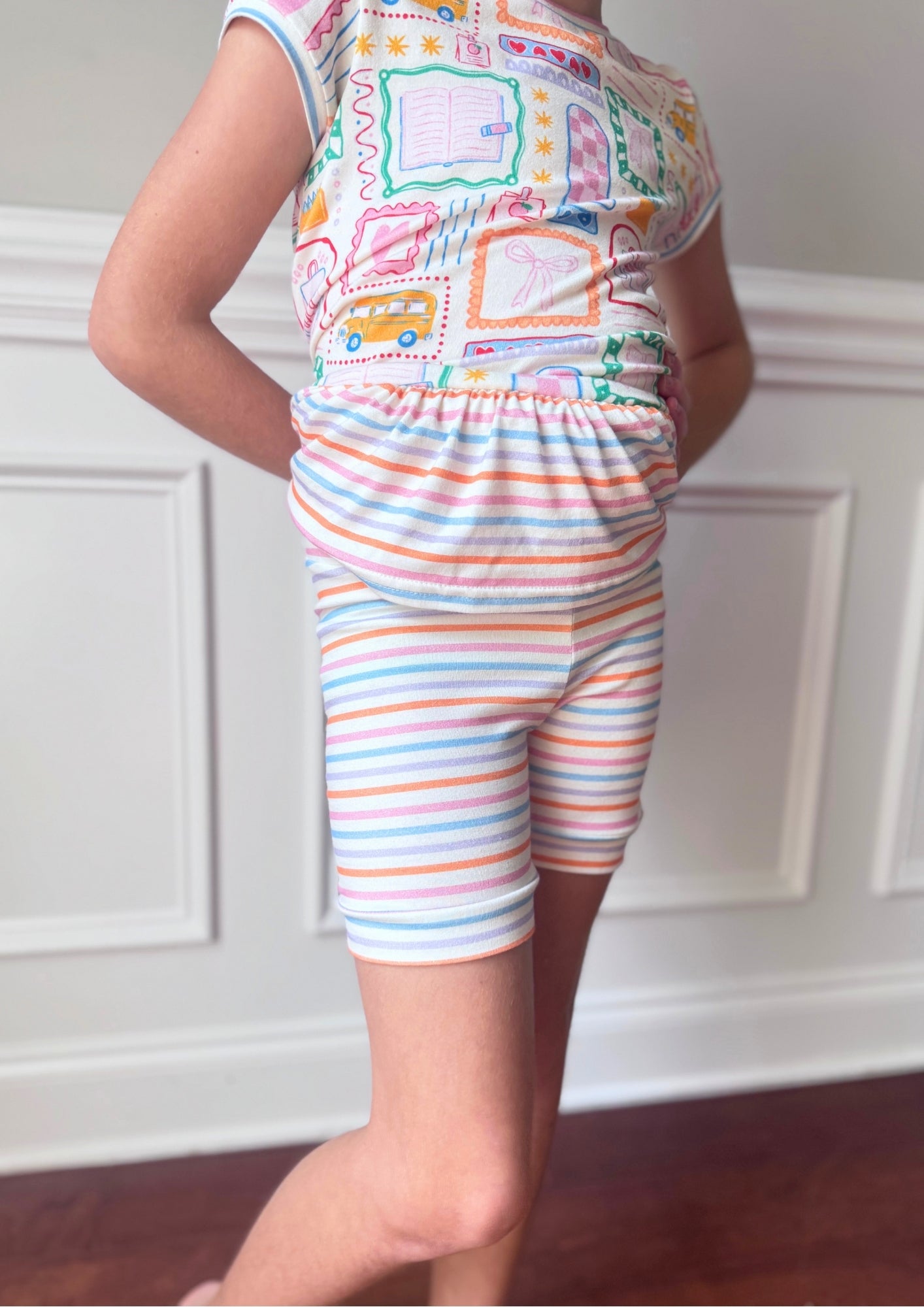 Child wearing a colorful outfit with a patterned top and striped shorts against a white wall.